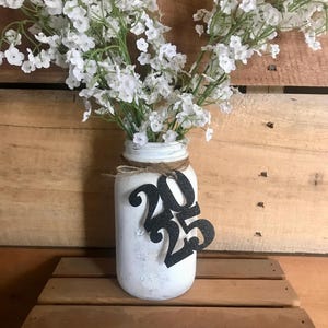 Puede incluir: Tarro de cristal blanco con un lazo de arpillera y un 2025 de purpurina negra sujeto al tarro. El tarro est&aacute; lleno de flores blancas de aliento de beb&eacute;.