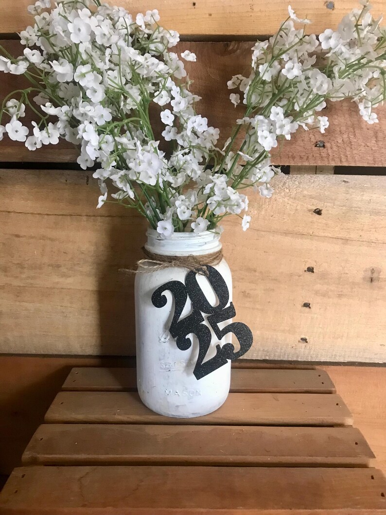 Puede incluir: Tarro de cristal blanco con un lazo de arpillera y un 2025 de purpurina negra sujeto al tarro. El tarro est&aacute; lleno de flores blancas de aliento de beb&eacute;.