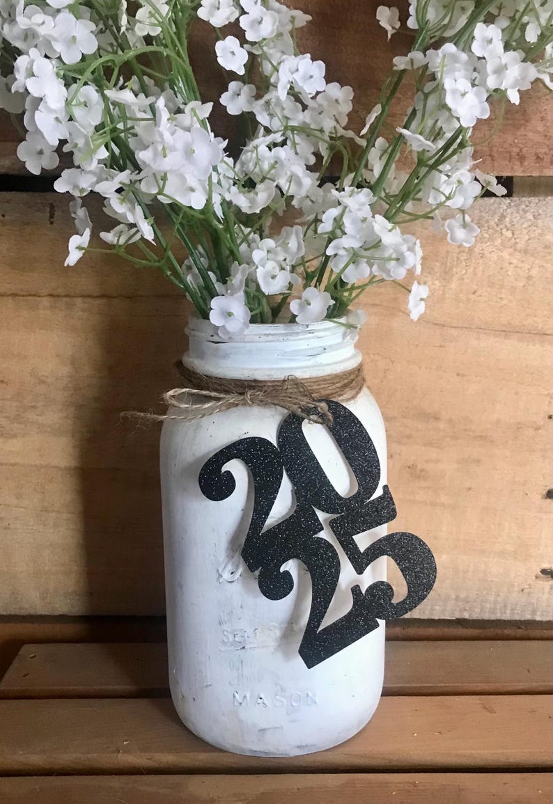 Puede incluir: Tarro de alba&ntilde;il blanco pintado con una cinta de arpillera y un recorte de purpurina negra 2023. El tarro est&aacute; lleno de flores blancas de aliento de beb&eacute;.