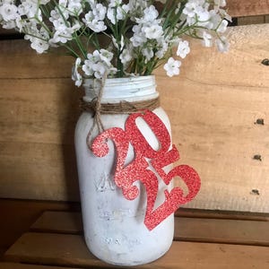 Puede incluir: Un tarro de alba&ntilde;il blanco pintado con una decoraci&oacute;n de purpurina roja 2023 y un ramo de aliento de beb&eacute; blanco.