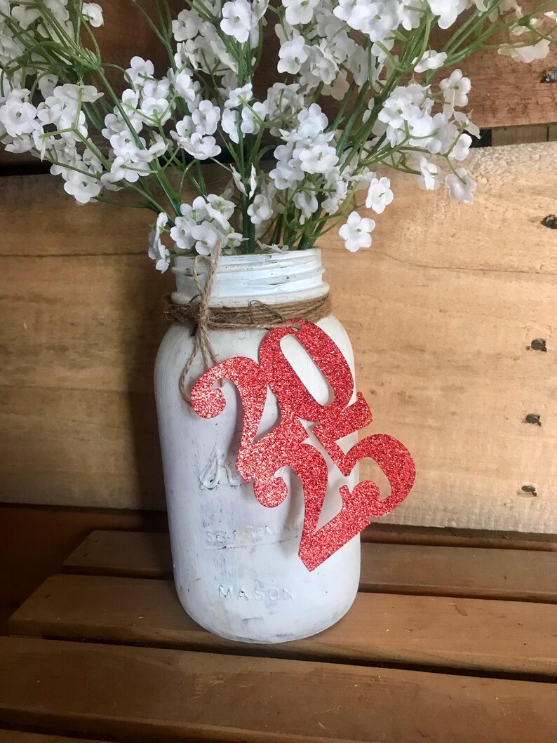 Puede incluir: Un tarro de alba&ntilde;il blanco pintado con una decoraci&oacute;n de purpurina roja 2023 y un ramo de aliento de beb&eacute; blanco.