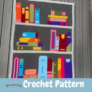 May include: A crochet pattern depicting a bookshelf design. The pattern shows three shelves with stacks of colourful books in various sizes and colours, including blue, orange, and pink. The text "Crochet Pattern" is visible at the bottom.