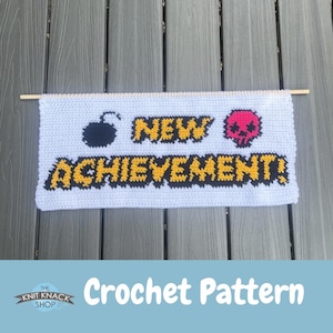 May include: A rectangular white crochet wall hanging suspended from a wooden dowel. The piece features the text NEW ACHIEVEMENT! in yellow with black outlines, flanked by a black bomb icon on the left and a pink skull icon on the right.