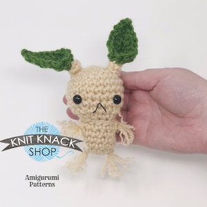 Puede incluir: Un juguete amigurumi de crochet con forma de planta, con un cuerpo de color marrón claro, hojas verdes y una carita pequeña y linda. El juguete está siendo sostenido por una mano.