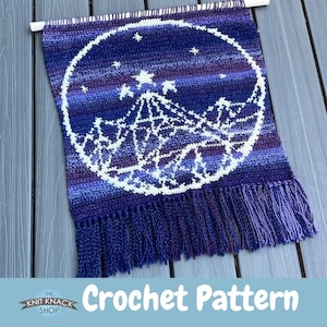Op de afbeelding: Een haakpatroon voor een wandhanger met een bergketen met sterren in een cirkel. Het ontwerp is uitgevoerd in tinten paars en blauw met een witte omtrek.