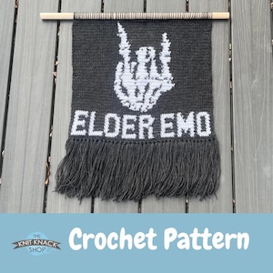 May include: A dark grey crochet wall hanging with the words "ELDER EMO" in white block letters. The design features a white hand gesture and fringe at the bottom. The wall hanging is attached to a wooden dowel.
