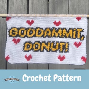 Może przedstawiać: Biała szydełkowa dekoracja ścienna z napisem "GODDAMMIT, DONUT!" w kolorze żółtym i czarnym, ozdobiona czerwonymi sercami. Znak jest przymocowany do drewnianego kołka. Słowa "Crochet Pattern" znajdują się na dole.