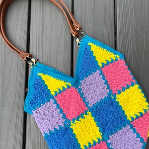 May include: A crocheted tote bag with a geometric pattern in shades of blue, yellow, pink, and purple. The bag has brown leather straps with metal clasps.