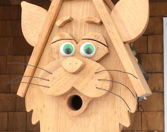 May include: A wooden birdhouse shaped like a cat's face with green eyes and a small opening for birds to enter. The birdhouse is hanging from a chain.