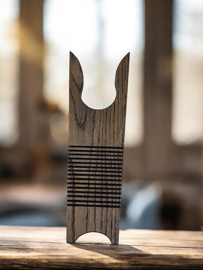 May include: A wooden weaving tool with a curved top and a series of horizontal black lines. The tool is made of light brown wood and has a smooth finish. The background is blurred, suggesting an indoor setting.