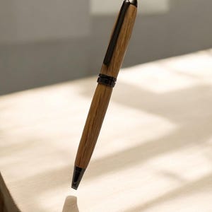 Pu&ograve; includere: Una penna in legno con clip e punta in metallo scuro. La penna ha un motivo a venature di legno marrone chiaro e un cappuccio in metallo nero. La penna &egrave; mostrata su uno sfondo chiaro.
