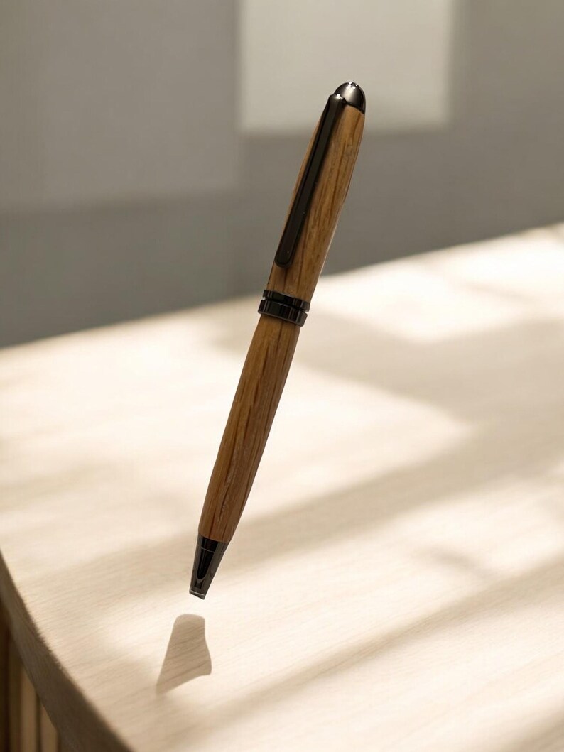 Pu&ograve; includere: Una penna in legno con clip e punta in metallo scuro. La penna ha un motivo a venature di legno marrone chiaro e un cappuccio in metallo nero. La penna &egrave; mostrata su uno sfondo chiaro.