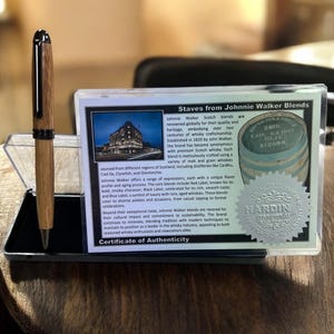 Pu&ograve; includere: Una penna in legno con dettagli argentati, esposta in una custodia trasparente. Accanto, un certificato di autenticit&agrave; per i blend di Scotch Johnnie Walker, con testo e un logo.