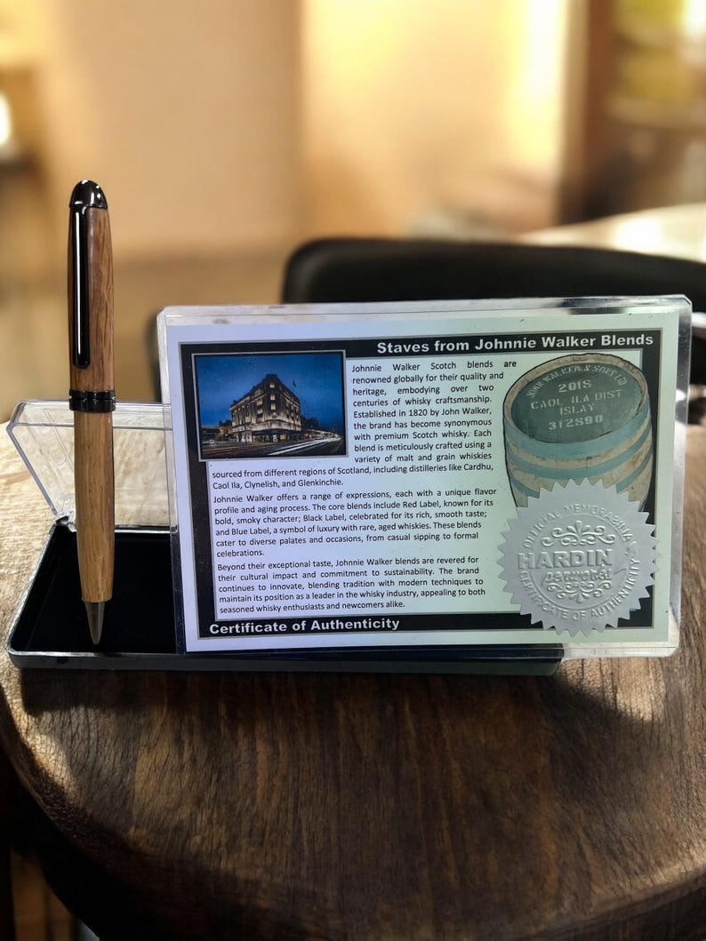 Pu&ograve; includere: Una penna in legno con dettagli argentati, esposta in una custodia trasparente. Accanto, un certificato di autenticit&agrave; per i blend di Scotch Johnnie Walker, con testo e un logo.