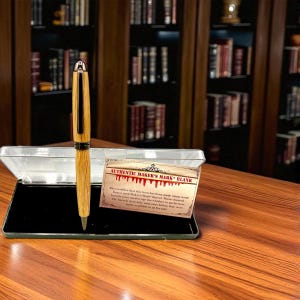May include: A wooden pen with a silver tip and accents, presented in a clear case with a black interior. The pen is made from light-colored wood with dark wood accents. A card reads "AUTHENTIC MAKER'S MARK BLANK".