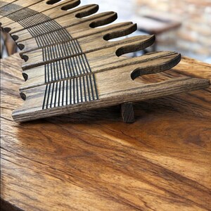 May include: A wooden book stand with a unique design. The stand is made of dark-stained wood and features a series of curved, finger-like supports. The supports are connected by horizontal slats with vertical grooves. The stand is on a wooden surface.