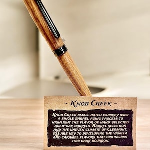 May include: A wooden pen with a black clip and accents, resting on a small card. The card features the text "~ KNOB CREEK -" and a description of the whiskey's aging process and flavor profile.