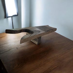 May include: A handcrafted wooden breadboard with a unique design. The board features a curved handle and a grooved section for slicing. The natural wood grain and warm tones create a rustic aesthetic. The breadboard is supported by a triangular base.