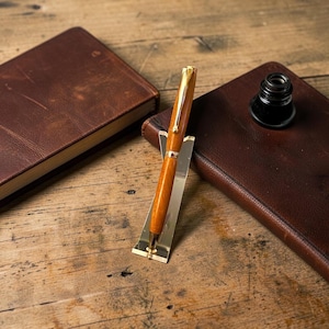 May include: A polished wooden pen with gold-toned metal accents, including a clip and center band, rests upright in a small, angled gold-colored metal stand. The pen features a smooth, light brown wood grain finish and a tapered, rounded shape.