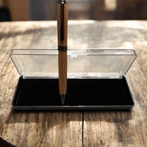 Pu&ograve; includere: Una penna in legno con punta e dettagli color argento, presentata in un astuccio di plastica trasparente con interno in velluto nero. La penna ha una finitura a venatura del legno marrone chiaro ed &egrave; su una superficie di legno.