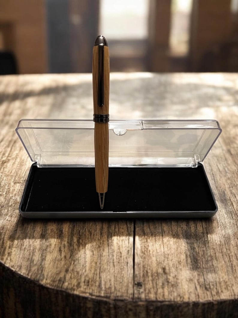 Pu&ograve; includere: Una penna in legno con punta e dettagli color argento, presentata in un astuccio di plastica trasparente con interno in velluto nero. La penna ha una finitura a venatura del legno marrone chiaro ed &egrave; su una superficie di legno.