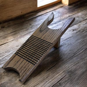 May include: A wooden boot jack with a light brown finish. The boot jack has a curved base, a grooved section, and a forked top. The boot jack is designed to help remove boots. The background is a wooden floor.
