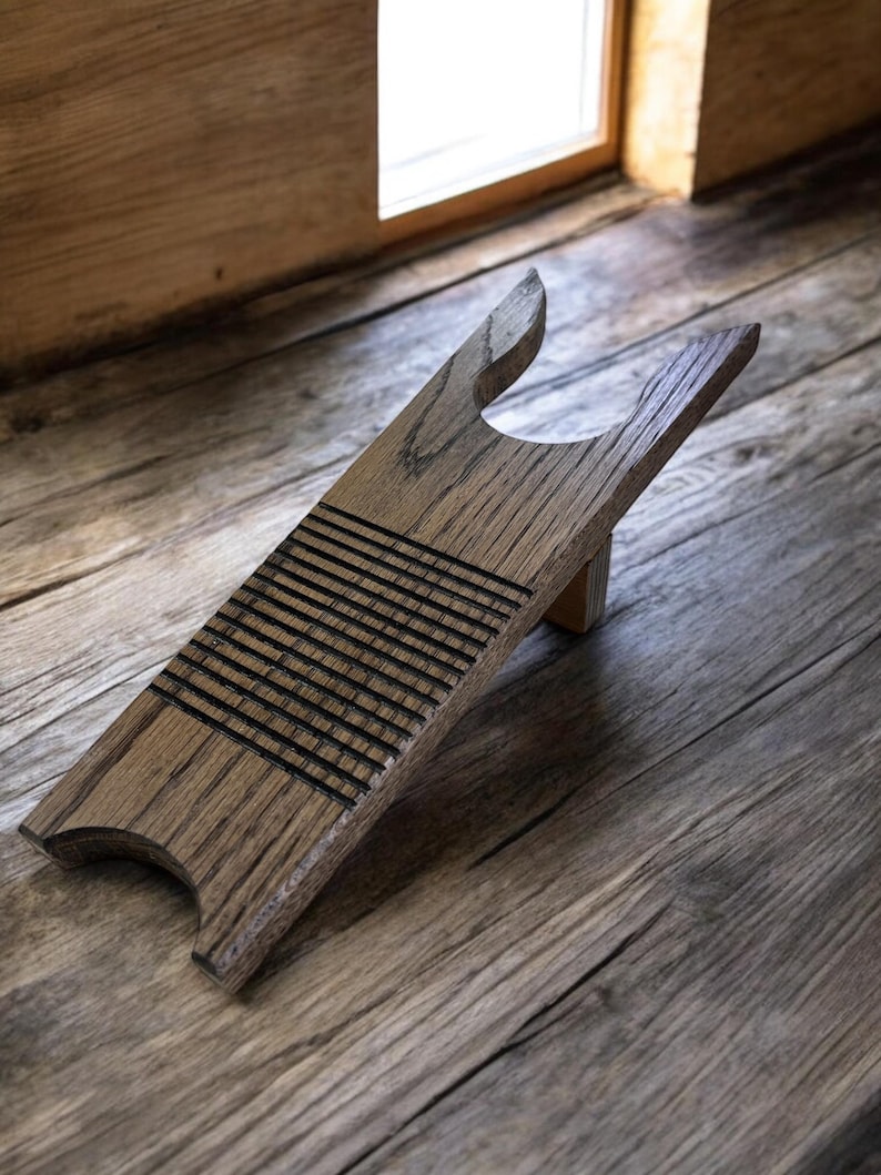 May include: A wooden boot jack with a light brown finish. The boot jack has a curved base, a grooved section, and a forked top. The boot jack is designed to help remove boots. The background is a wooden floor.
