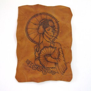 Op de afbeelding: Een handgemaakt leren kunstwerk met een geisha met een paraplu en een waaier. De geisha is in zwarte lijnen op het bruine leer getekend. Kersenbloesems zijn aan de onderkant geëtst. Het leer heeft een natuurlijke, onregelmatige rand.