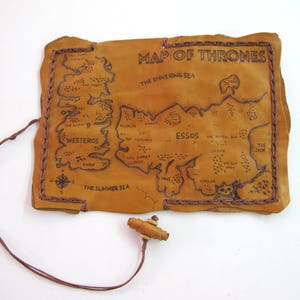 May include: A tan leather map of the fictional world from the series "Game of Thrones", with the title "Map of Thrones" at the top. The map is hand-drawn with black ink and features the continents of Westeros and Essos. A brown leather cord and wooden toggle are attached.