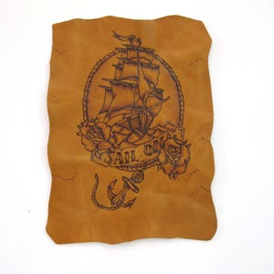 May include: A tan leather piece with a detailed black illustration of a sailing ship, roses, and an anchor. The words "SAIL ON" are displayed on a banner. The leather has a distressed, aged appearance.