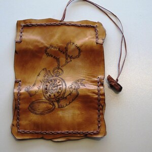 May include: A rectangular, tan leather pouch with a brown stitched border and a brown cord. The pouch features a detailed illustration of a pocket watch with a chain and a decorative border. The leather has a distressed, aged appearance.