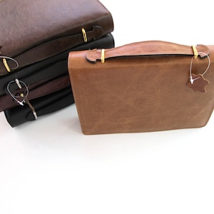 May include: A stack of four leather-bound journals with gold clasps and leather tags. The top journal is brown, the next two are black, and the bottom journal is brown.
