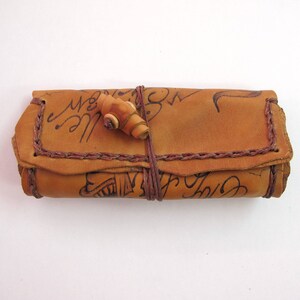 May include: A rolled tan leather case with a wooden clasp. The case features dark brown stitching and handwritten script. The leather has a worn appearance. It is secured with a leather cord. The case is likely for storing small items.