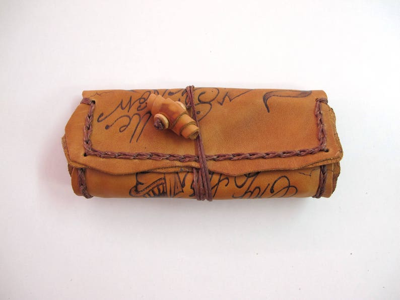 May include: A rolled tan leather case with a wooden clasp. The case features dark brown stitching and handwritten script. The leather has a worn appearance. It is secured with a leather cord. The case is likely for storing small items.