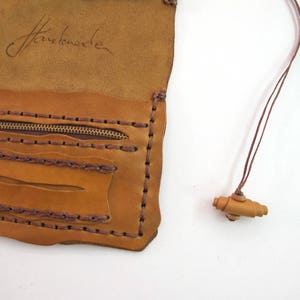 May include: Tan and brown leather pouch with a zip pocket and a small wooden toggle on a cord. The pouch is handmade, with visible stitching and a handwritten signature. The measurements are in centimetres.