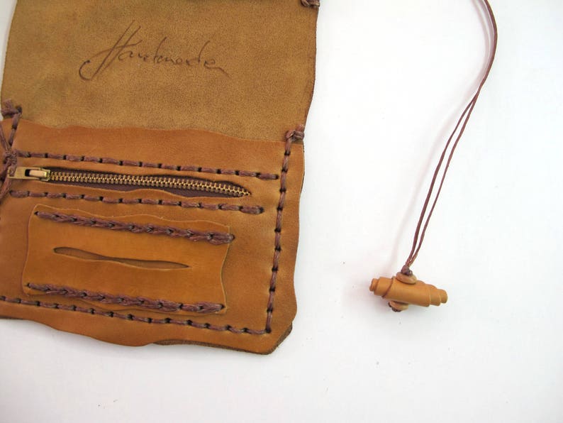 May include: Tan and brown leather pouch with a zip pocket and a small wooden toggle on a cord. The pouch is handmade, with visible stitching and a handwritten signature. The measurements are in centimetres.