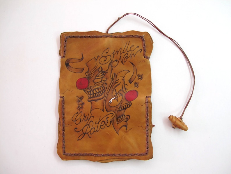 May include: A tan leather pouch with a brown stitched border and a brown cord. The pouch features a black illustration of two clown faces with red noses and the words "Smile Now, Cry Later."