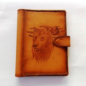 May include: A tan leather wallet with a detailed embossed image of a wolf's head and a sunset scene. The wallet has a snap closure and stitched edges. The overall design is rustic and handcrafted.