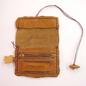 May include: Tan leather wallet, handmade with dark brown stitching. It has a zip pocket, a small rectangular pocket, and a flap closure. The word 'Handmade' is inscribed. A brown leather cord with a wooden bead is attached.