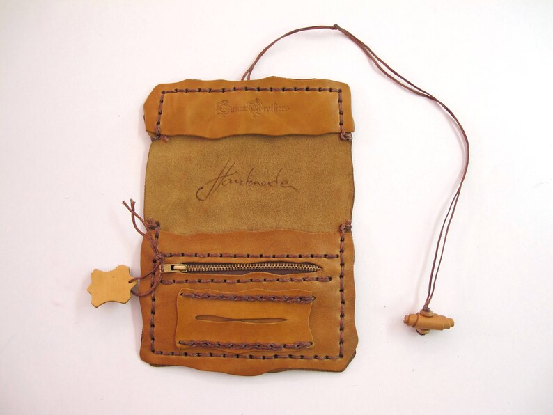 May include: Tan leather wallet, handmade with dark brown stitching. It has a zip pocket, a small rectangular pocket, and a flap closure. The word 'Handmade' is inscribed. A brown leather cord with a wooden bead is attached.