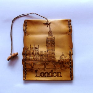 May include: A tan leather wall hanging featuring a detailed depiction of London's Big Ben and the Houses of Parliament. The word "London" is etched at the bottom. A wooden toggle and cord are attached at the top.