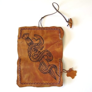 May include: Handmade leather pouch with a dagger and snake design. The brown pouch has a stitched border and a leather cord with a wooden bead. A small leather tag hangs from the bottom.