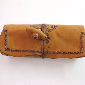 May include: A handcrafted leather case in a warm tan color, featuring a stitched border and a decorative butterfly design. The case is secured with a wooden toggle and a leather strap, showcasing a rustic aesthetic.