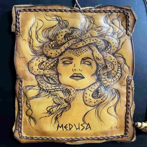 May include: Hand-tooled leather artwork featuring Medusa with snakes for hair. The artwork is in shades of tan and brown, with the word "MEDUSA" at the bottom. The edges are stitched with a braided border.