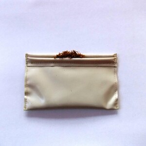May include: A cream-coloured, rectangular pouch with a folded top, filled with loose, brown tobacco. The pouch is made of a smooth, possibly leather-like material. The background is plain white.