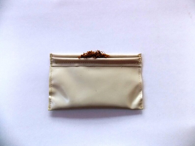 May include: A cream-coloured, rectangular pouch with a folded top, filled with loose, brown tobacco. The pouch is made of a smooth, possibly leather-like material. The background is plain white.