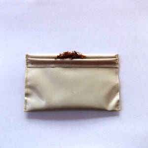 May include: A cream-colored, rectangular pouch with a folded top, filled with loose, brown tobacco. The pouch has a stitched border and a slightly glossy finish. The background is plain white.