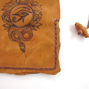 May include: Tan leather pouch with a hand-drawn design of an eye within a circle, surrounded by a snake and floral elements. The edges are stitched with a darker brown thread. A small wooden toggle is attached.