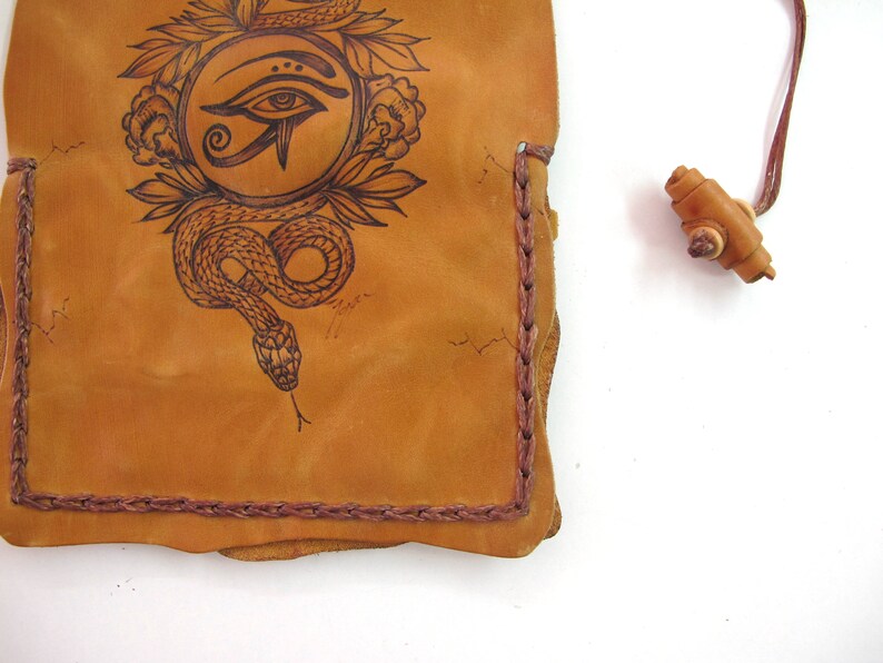 May include: Tan leather pouch with a hand-drawn design of an eye within a circle, surrounded by a snake and floral elements. The edges are stitched with a darker brown thread. A small wooden toggle is attached.