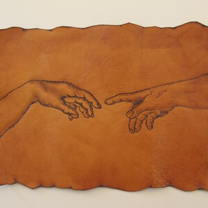 May include: A rectangular, tan leather piece with a dark brown, hand-drawn depiction of two hands nearly touching, reminiscent of Michelangelo's "The Creation of Adam." The edges of the leather are uneven, adding a rustic feel.